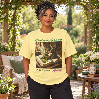 A woman wearing a pale yellow graphic t-shirt in a garden patio setting. The t-shirt features a vintage-style illustration of a man proposing to a fairy as they sit on an open book, with the text 'I found my happily ever after in the pages of a Romantasy'.
