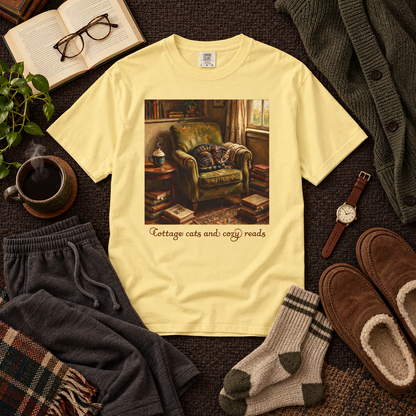 A pale yellow graphic T-shirt laid flat on a dark textured surface, surrounded by cozy masculine props including folded sweatpants, slippers, socks, a mug of coffee, an open book, glasses, a cardigan, and a watch. The shirt features a printed illustration of a cozy armchair scene with the text 'Cottage cats and cozy reads'.