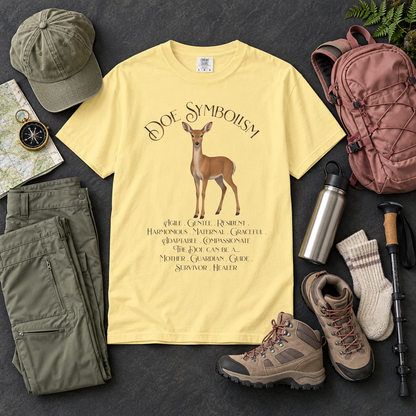 A pale yellow graphic T-shirt laid flat on a dark textured surface, surrounded by outdoor gear including an olive green cap, matching cargo pants, hiking boots, a pink backpack, a metal water bottle, a trekking pole, and a pair of knit socks. The shirt features a deer illustration and the text 'Doe Symbolism' with a list of meanings associated with the female deer.