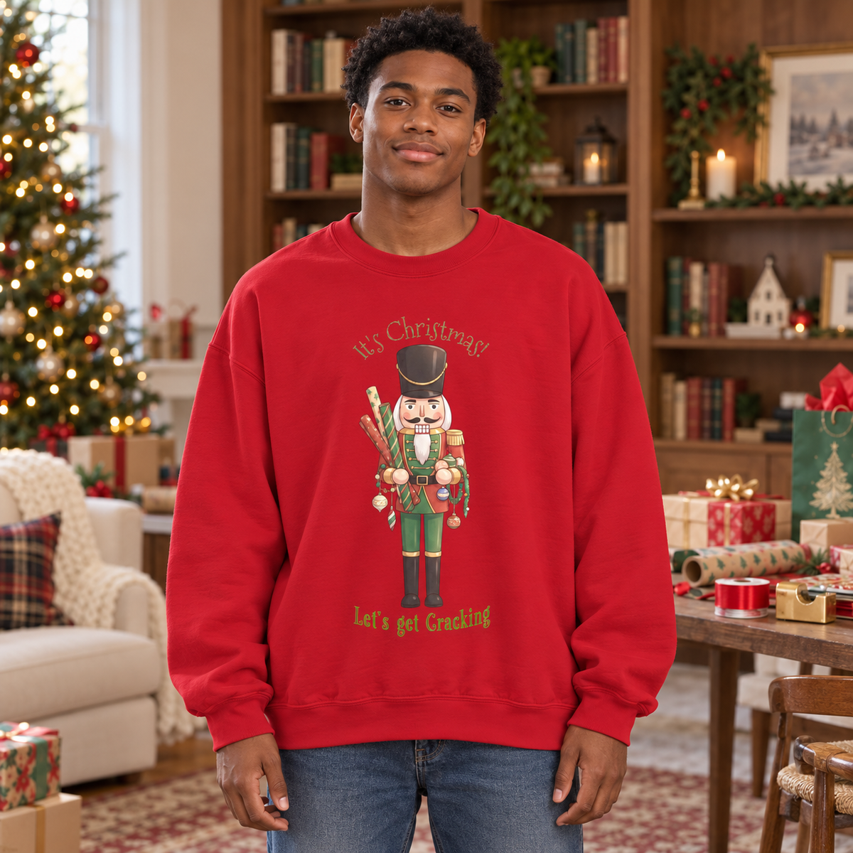 A man wearing a red crewneck sweatshirt with a nutcracker graphic and the text 'It's Christmas!' and 'Let's get Cracking'. The background shows a cozy, decorated Christmas living room with a tree, bookshelves, gift bags, wrapping paper, ribbons, tape, and gifts.