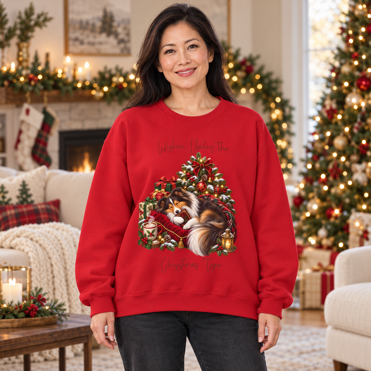 A woman wearing a red crewneck sweatshirt with an illustration of a sheltie sleeping under a festive Christmas tree and the text 'Wishin' Under the Christmas Tree'. The woman stands in a decorated living room with a Christmas tree, stockings, and a fireplace.