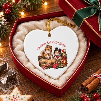 A heart-shaped white ceramic Christmas ornament with a gold hanging loop, featuring an illustration of two shelties at a holiday table and the text 'Christmas Bakes Life Better.' The ornament is displayed inside a red gift box with cream cushioning and a green ribbon on a rustic wooden table decorated with pine branches, lights, cookies, cinnamon sticks, and cookie cutters.
