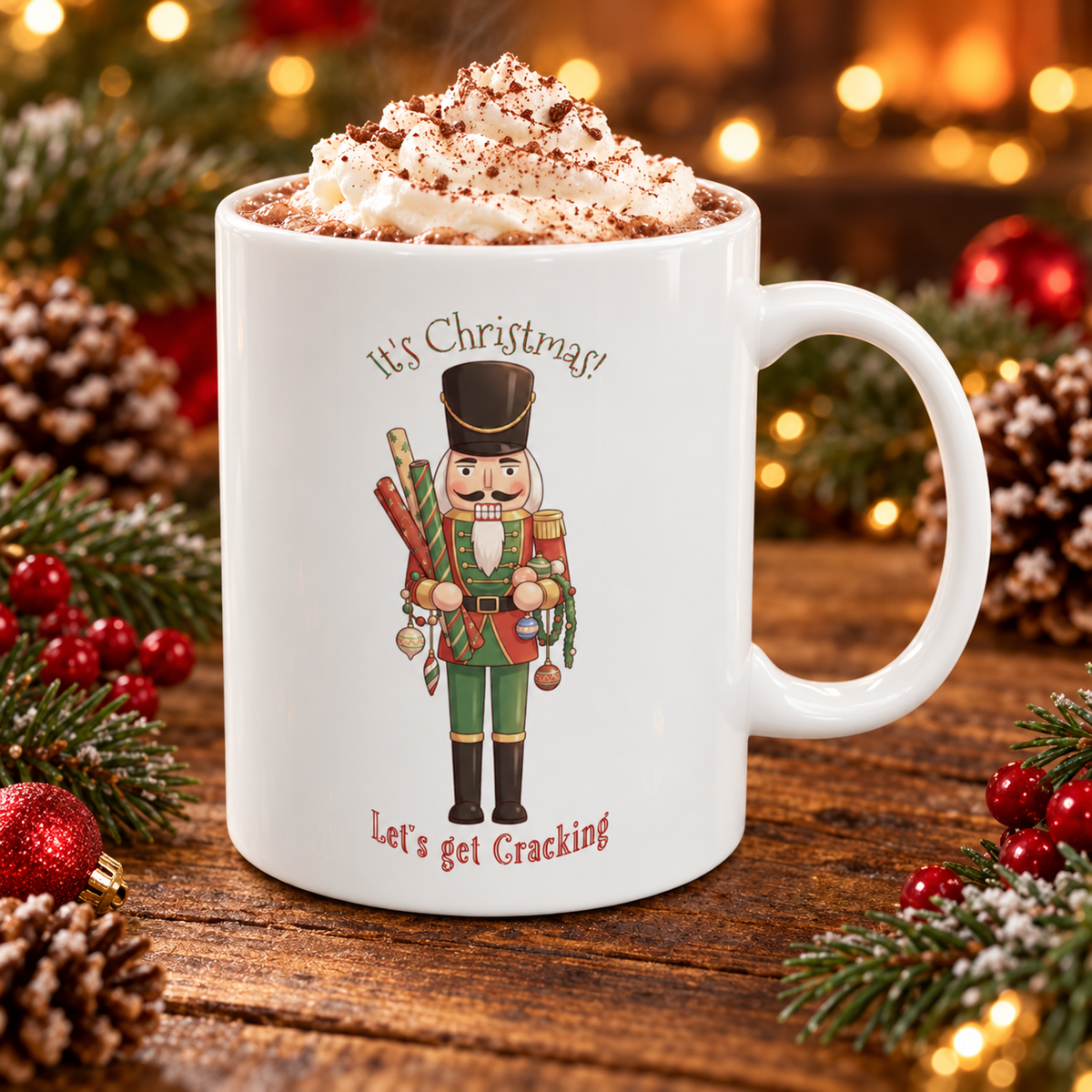 A white ceramic mug filled with whipped cream and cocoa, decorated with a nutcracker illustration and the text 'It's Christmas' and 'Let's get Cracking'. The mug is set on a wooden surface surrounded by pine branches, red berries, pinecones, and warm holiday lights.