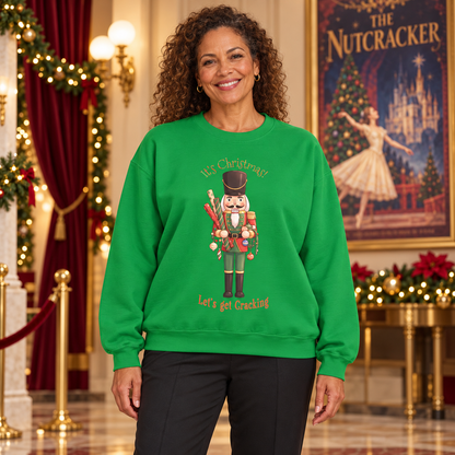 A woman wearing a bright green crewneck sweatshirt with a nutcracker graphic and the text 'It's Christmas! Let's get Cracking' stands in a decorated holiday interior with red drapery, lights, and a framed 'The Nutcracker' poster in the background.