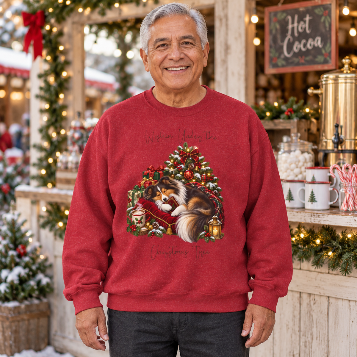 A smiling older man wearing a red crewneck sweatshirt with a Christmas-themed Shetland Sheepdog sleeping under a Christmas tree and the words, 'Wishin' Under the Christmas Tree'. The man is standing in front of a festive outdoor hot cocoa stall that is decorated with lights and holiday greenery.