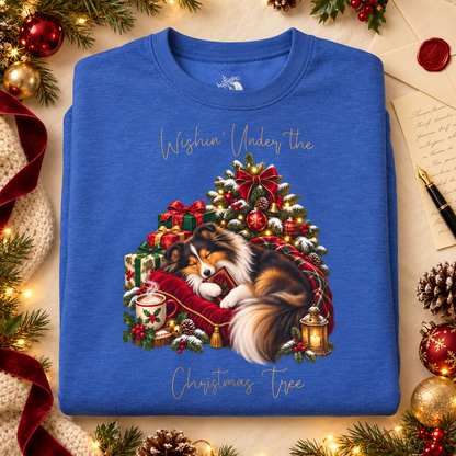 A folded flat-lay of a heathered royal blue sweatshirt with an illustration of a Shetland sheepdog curled under a Christmas tree and the words, 'Wishin' Under the Christmas tree'. Surrounding the shirt are ornaments, a letter, and a pen.