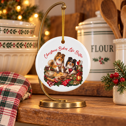 Round white ceramic Christmas ornament displayed on a gold stand on a wooden table with kitchen canisters, bowls, a plaid cloth, and holiday decor in the background. The ornament features an illustration of two shelties baking with the text 'Christmas Bakes Life Better.'