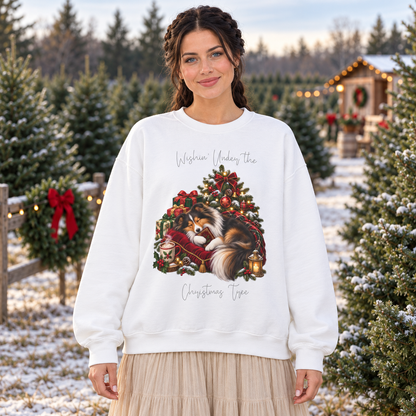 A woman stands outdoors in a snowy Christmas tree lot wearing a white crewneck sweatshirt with a large graphic of a collie dog wrapped sleeping near a Christmas wreath, surrounded by gifts and a steaming mug. The text on the sweatshirt reads, 'Wishin' Under the' above the graphic and 'Christmas Tree' below it.