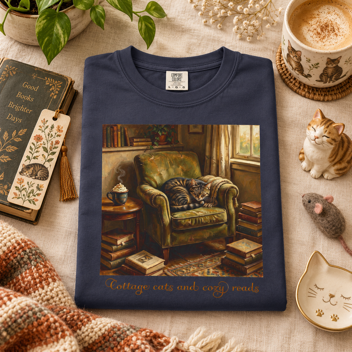 A navy blue crewneck t-shirt is folded and presented in a cozy flat-lay setting. The shirt features a large graphic print of a cozy scene with a cat sleeping in an armchair, surrounded by books, with the text 'Cottage cats and cozy reads' printed below.