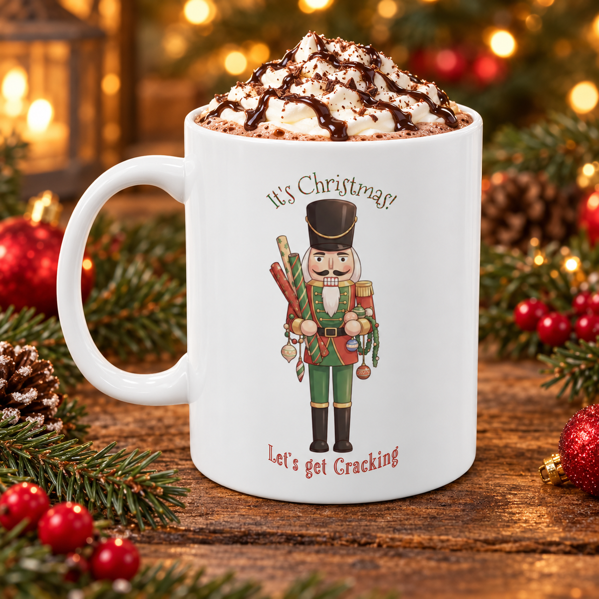 A white ceramic mug filled with a whipped hot chocolate drink topped with chocolate drizzle, set on a wooden surface with Christmas greenery and ornaments around it. The mug features a nutcracker illustration with the text 'It's Christmas! Let's Get Cracking'.