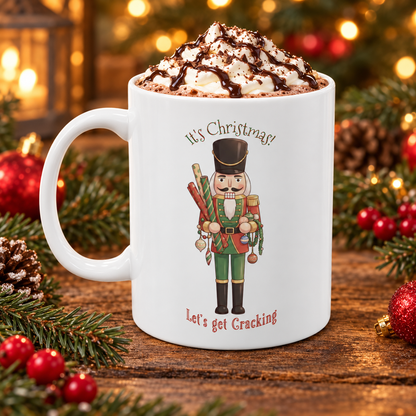 A white ceramic mug filled with a whipped hot chocolate drink topped with chocolate drizzle, set on a wooden surface with Christmas greenery and ornaments around it. The mug features a nutcracker illustration with the text 'It's Christmas! Let's Get Cracking'.