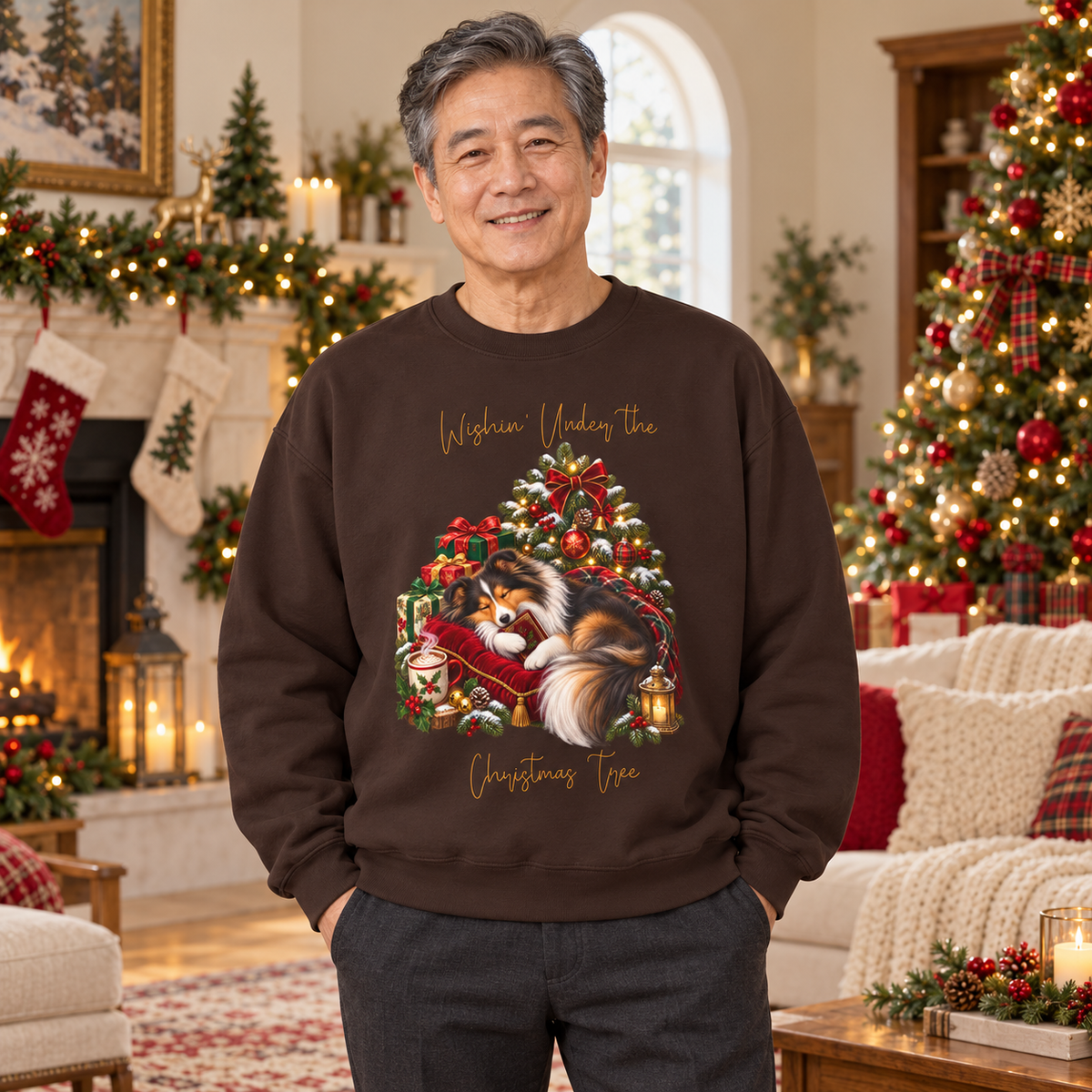 A smiling man stands in a decorated Christmas living room wearing a dark brown crewneck sweatshirt with a festive graphic of a fluffy dog curled up on a red velvet cushion, surrounded by a glowing Christmas tree and the words 'Wishin' Under the Christmas Tree'.