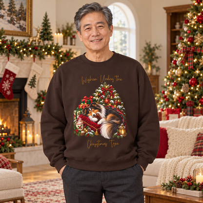 A smiling man stands in a decorated Christmas living room wearing a dark brown crewneck sweatshirt with a festive graphic of a fluffy dog curled up on a red velvet cushion, surrounded by a glowing Christmas tree and the words 'Wishin' Under the Christmas Tree'.