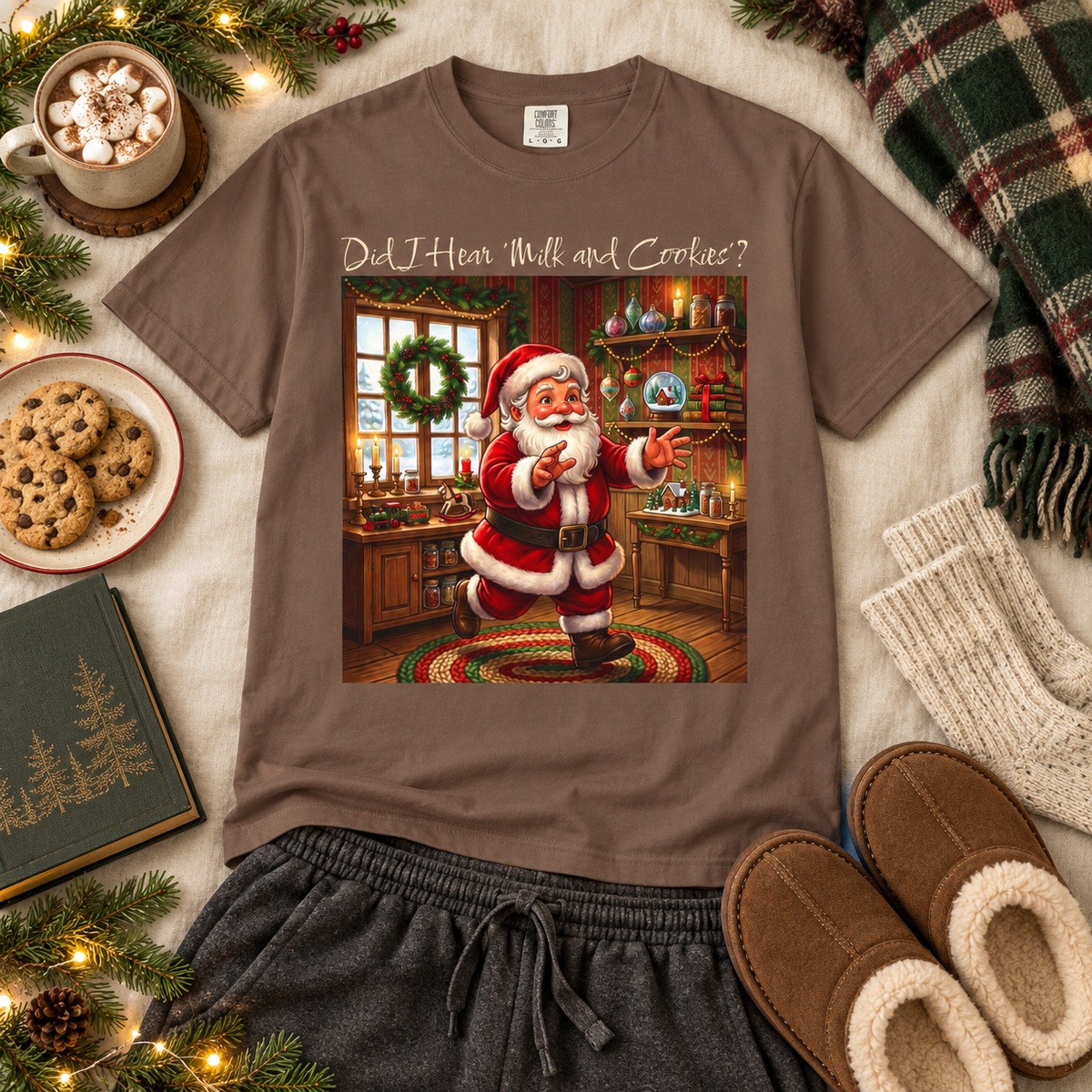 A brown short-sleeve graphic T-shirt laid flat on a cozy holiday-themed surface with a mug of hot chocolate, cookies, knit socks, slippers, a book, and pine branches. The shirt has a large front print of Santa Claus in a decorated workshop with the text "Did I Hear 'Milk and Cookies'?".