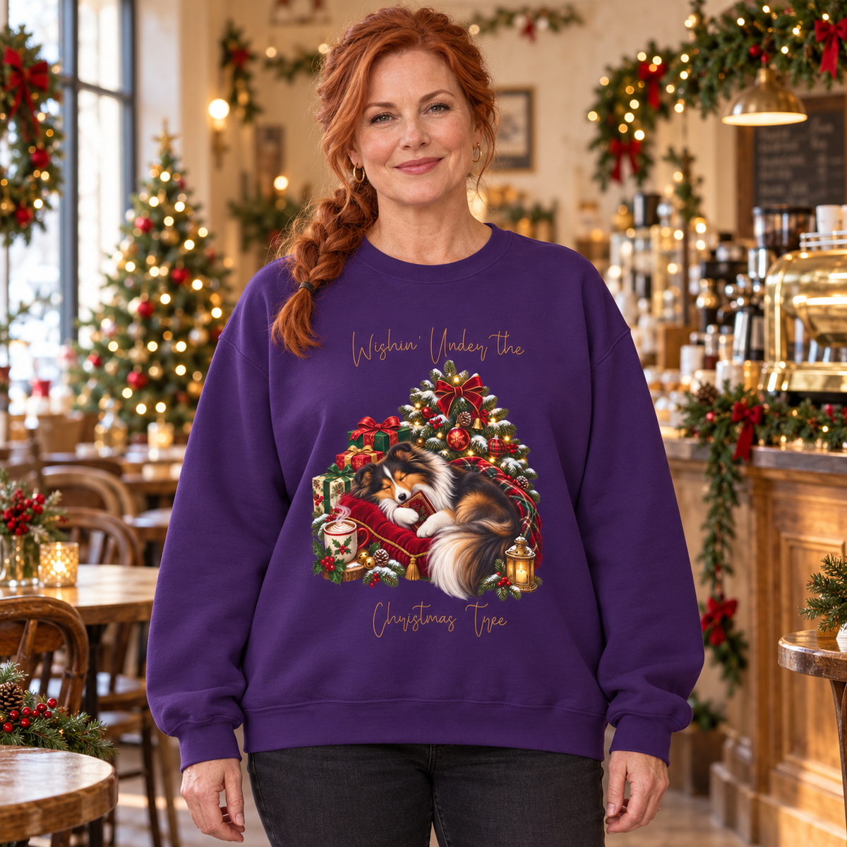A woman wearing a purple crewneck sweatshirt with a sheltie curled under a Christmas tree and the text 'Wishin' Under the Christmas Tree'. The woman is standing in a decorated holiday cafe.