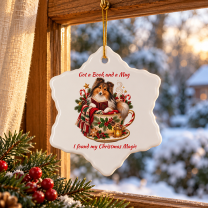 A white ceramic Christmas ornament shaped like a snowflake hangs from a wooden window frame. The ornament features an illustration of a Sheltie reading a book in a teacup surrounded by holiday decorations, with red text reading 'Got a Book and a Mug' and 'I found my Christmas Magic'.
