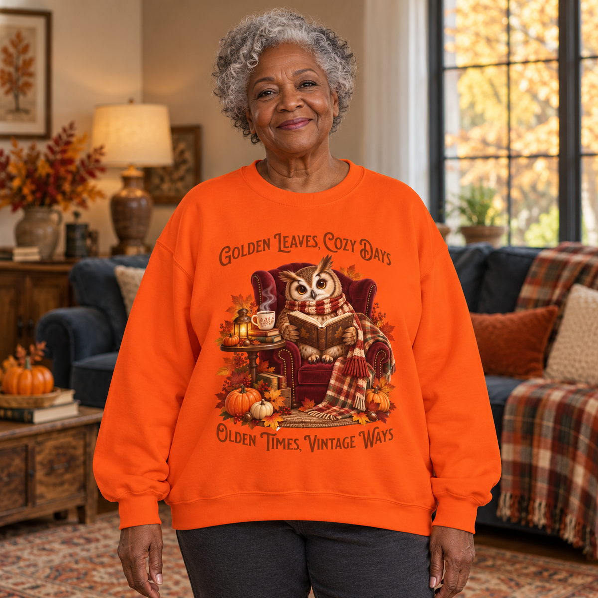 An older woman stands in a cozy living room wearing a bright orange crewneck sweatshirt with a large fall-themed owl graphic and the text 'Golden Leaves, Cozy Days' and 'Olden Times Vintage Ways.'.