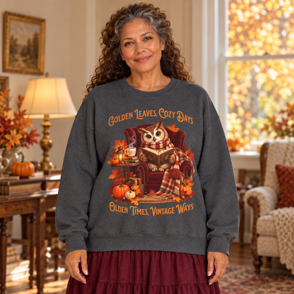 A woman wearing a dark gray crewneck sweatshirt with a cozy autumn owl graphic and the text 'Golden Leaves, Cozy Days' and 'Golden Times, Vintage Ways', posed in a warm, decorated living room.