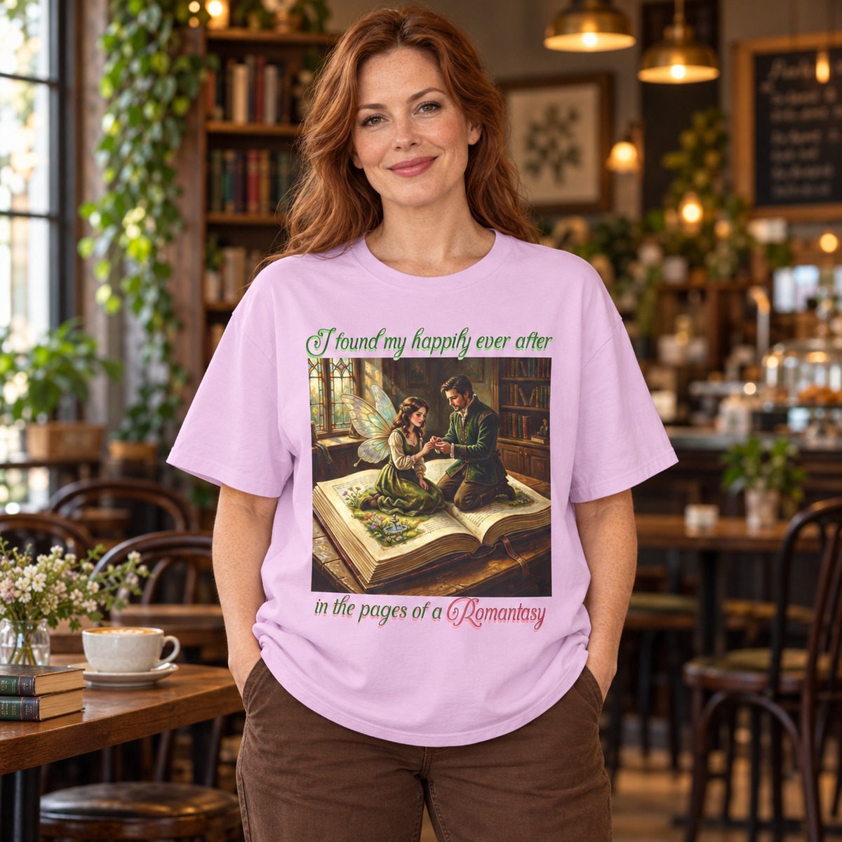 A woman stands in a cozy, softly blurred cafe interior wearing a light lavender graphic t-shirt. The t-shirt features a central illustration of a man proposing to a fairy as they sit on an open book, with the text 'I found my happily ever after in the pages of a Romantasy' in green and pink lettering.