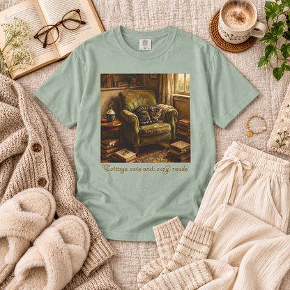A mint green crewneck t-shirt featuring a cozy reading room graphic with a green armchair and the text 'Cottage cats and cozy reads'. The shirt is styled in a flat lay with a beige knit cardigan, a woman's slippers, cream lounge pants, a cup of coffee, glasses, and an open book.