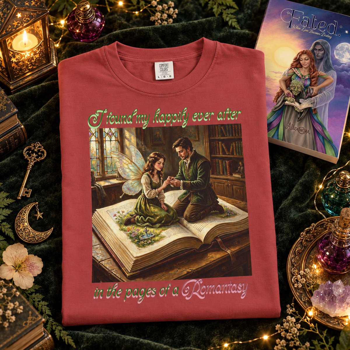 A folded red graphic T-shirt displayed on a dark green velvet surface with fantasy-themed props, including a book called Fated, key, candle, and flowers. The shirt features a romantic illustration of a man proposing to a fairy as they sit on a large open book, with decorative text reading 'I found my happily ever after in the pages of a Romantasy'.