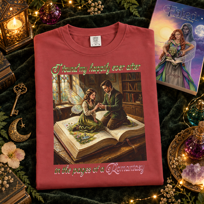 A folded red graphic T-shirt displayed on a dark green velvet surface with fantasy-themed props, including a book called Fated, key, candle, and flowers. The shirt features a romantic illustration of a man proposing to a fairy as they sit on a large open book, with decorative text reading 'I found my happily ever after in the pages of a Romantasy'.
