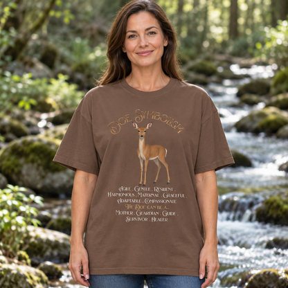 A woman stands outdoors in front of a flowing stream in a forest, wearing a brown graphic T-shirt with a deer illustration and the text 'Doe Symbolism' on the front.