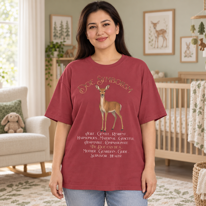 A woman modeling a short-sleeve maroon graphic t-shirt in a cozy nursery setting. The shirt features a central deer graphic and the text 'Doe Symbolism' along with a list of positive qualities.