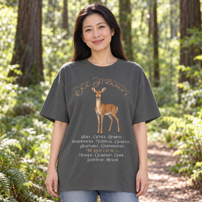 A woman standing in a sunlit forest wearing an oversized dark charcoal gray t-shirt with a graphic of a deer and the text 'Doe Symbolism' and a list of qualities. She is also wearing blue jeans.