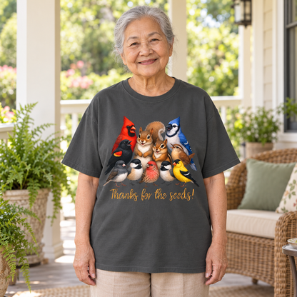 An older woman stands on a porch wearing a dark charcoal gray t-shirt with a colorful graphic of various birds, a squirrel, and a chipmunk and the text 'Thanks for the seeds!'