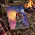 A book titled "Fated: When Love Becomes Destiny" is placed on a mossy rock outdoors near a campfire, with a blanket and coffee mug visible at the left edge. The cover art shows semi transparent man standing behind a woman who is grabbing the quill of a green gremlin and attempting to pour a drink on the gremlin's open book. The man has his hand under the liquid. Behind them is a full moon and stars on the right and on the left a bright sun.