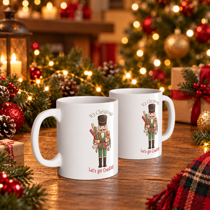 Two white ceramic Christmas mugs with nutcracker illustrations and the phrases 'It's Christmas' and 'Let's get cracking!' displayed on a wooden table in a festive holiday setting with lights, ornaments, and pine branches.