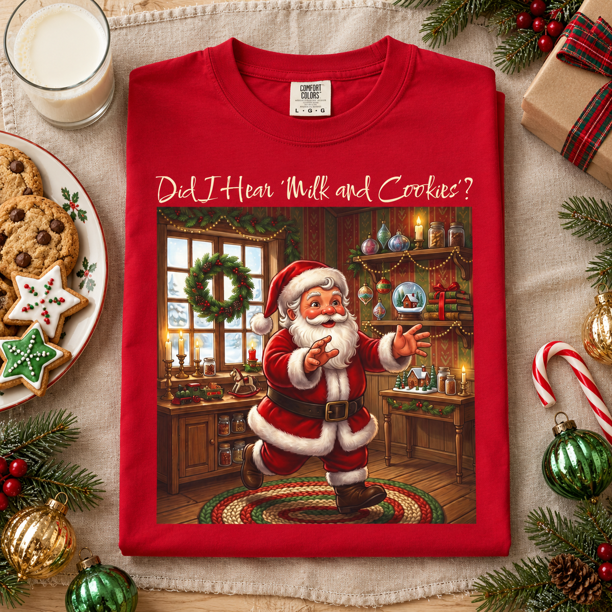 A red crewneck t-shirt featuring a festive graphic of a running Santa Claus in a decorated holiday room, with the text 'Did I Hear "Milk and Cookies"?' printed above him. The shirt is displayed in a styled flat-lay setting surrounded by Christmas decorations, cookies, a glass of milk, and a wrapped gift on a beige fabric background.