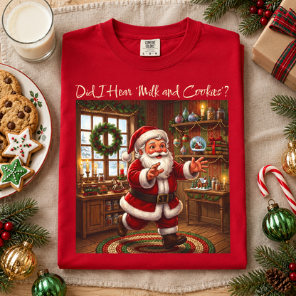 A red crewneck t-shirt featuring a festive graphic of a running Santa Claus in a decorated holiday room, with the text 'Did I Hear "Milk and Cookies"?' printed above him. The shirt is displayed in a styled flat-lay setting surrounded by Christmas decorations, cookies, a glass of milk, and a wrapped gift on a beige fabric background.