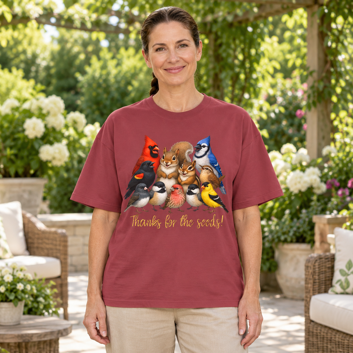 A woman wearing a maroon short-sleeve t-shirt with a colorful graphic of various birds, a squirrel, and a chipmunk and the text 'Thanks for the seeds!' printed on the front. She stands in an outdoor patio setting with wicker furniture and greenery.