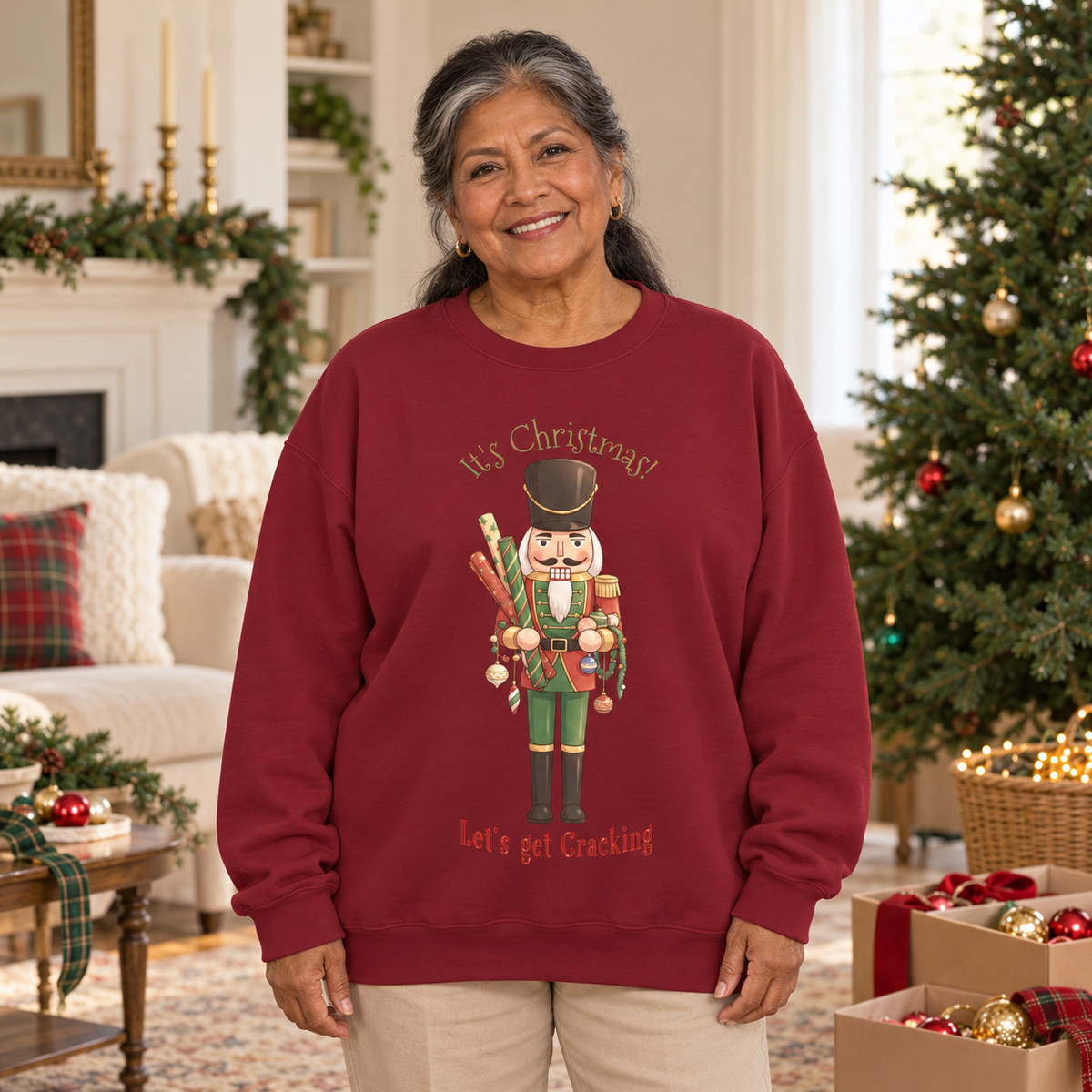 A smiling older woman stands in a decorated living room wearing a burgundy sweatshirt with a nutcracker holding decorations and wrapping paper and the text 'It's Christmas' and 'Let's get Cracking'.