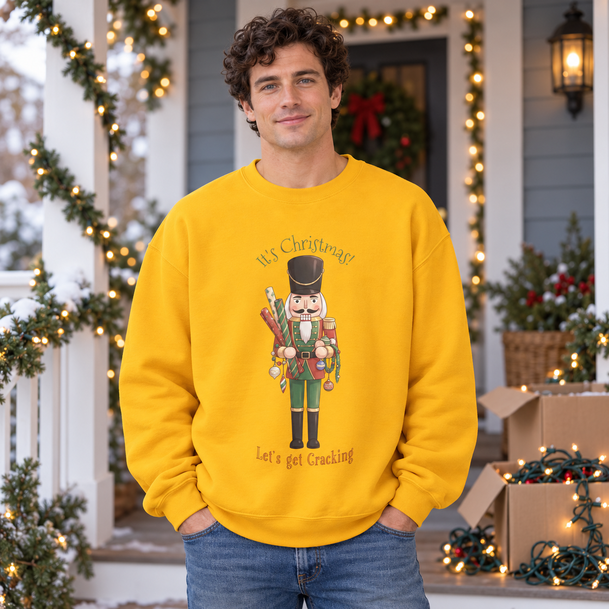 A man wearing a bright yellow crewneck sweatshirt with a Christmas nutcracker holding decorations and gift wrap surrounded by the text 'It's Christmas! Let's Get Cracking'. The man stands on a decorated holiday porch with boxes of lights.