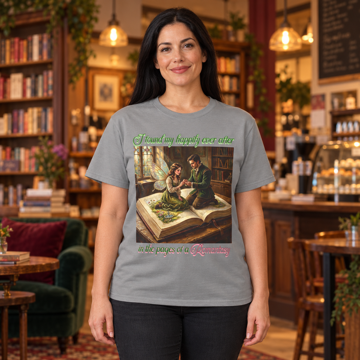 A woman wearing a light gray graphic t-shirt with a vintage-style illustration of a fairy and a man sitting on an open book. The woman stands in front of a cozy, blurred bookshop's cafe.