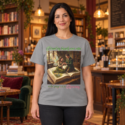 A woman wearing a light gray graphic t-shirt with a vintage-style illustration of a fairy and a man sitting on an open book. The woman stands in front of a cozy, blurred bookshop's cafe.