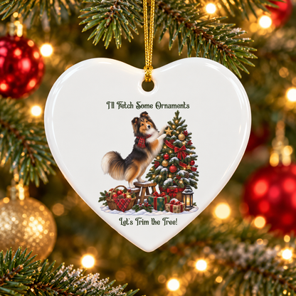 A heart-shaped white ceramic Christmas ornament hanging from a gold string on a decorated Christmas tree. The ornament features an illustration of a sheltie standing on a step stool next to a Christmas tree with ornaments and wrapped gifts, with the text 'I'll Fetch Some Ornaments' above and 'Let's Trim the Tree!' below.