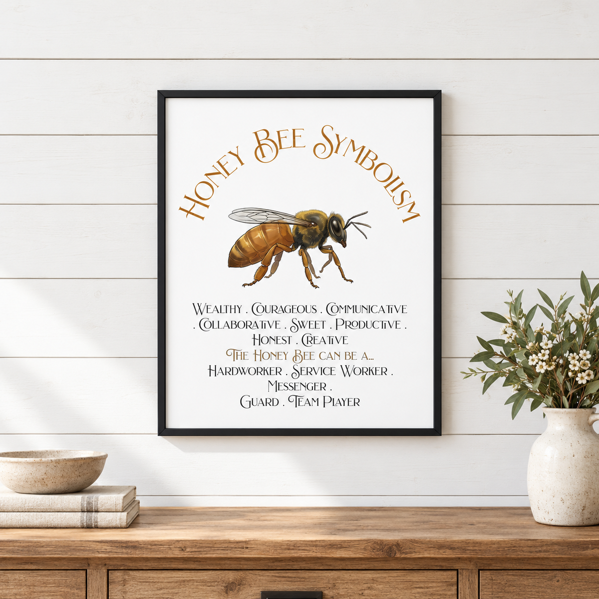A framed art print featuring a honey bee illustration and the text 'Honey Bee Symbolism' with symbolic text, displayed on a white shiplap wall above a wooden console table with a vase of flowers, a bowl, and books.
