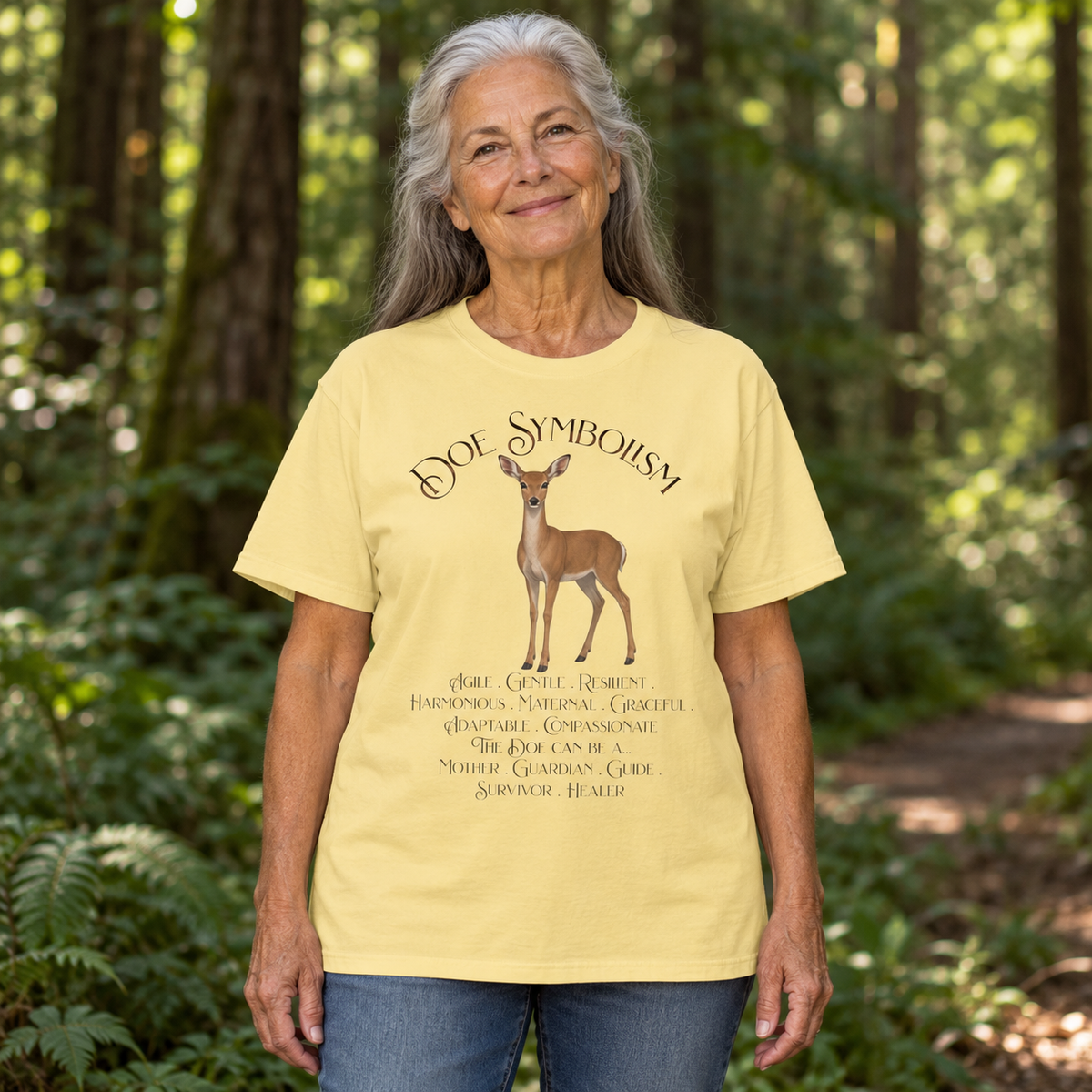 An older woman with gray hair stands in a sun-dappled forest . She wears a pale yellow graphic T-shirt with a deer illustration and the text 'DOE SYMBOLISM' and descriptive script text below.