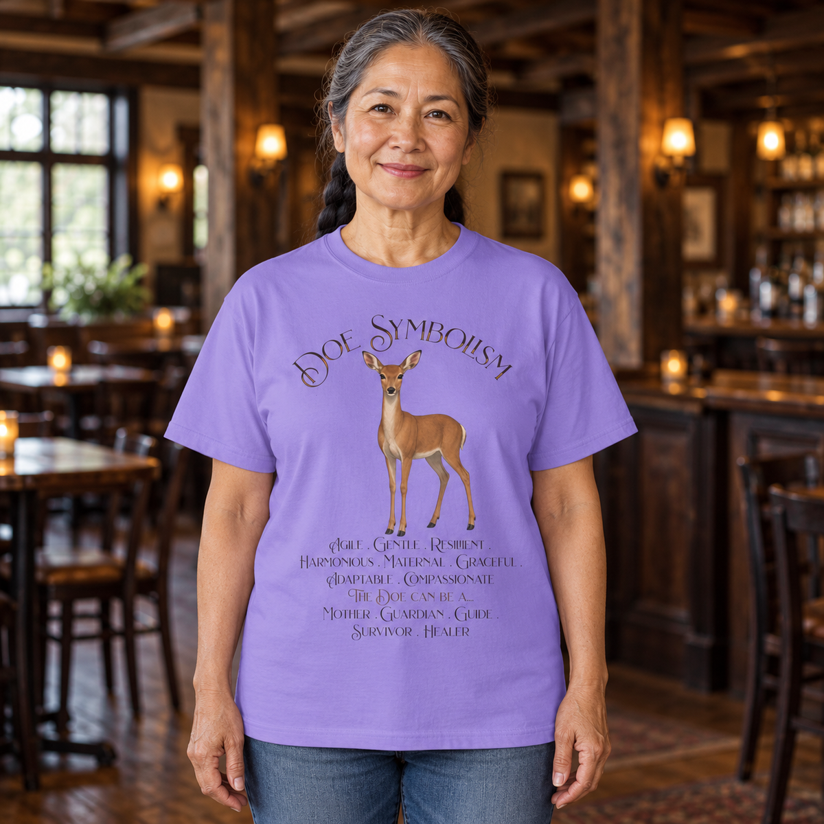 A woman wearing a lavender purple t-shirt with a deer graphic and Doe Symbolism title. She stands in a rustic wooden tavern interior.