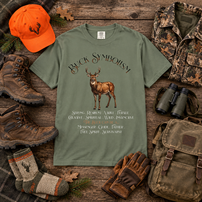 An olive green graphic t-shirt featuring a deer illustration and the text 'Buck Symbolism' surrounded by hunting-themed props like an orange cap, boots, binoculars, gloves, socks, and a backpack on a rustic wood background.