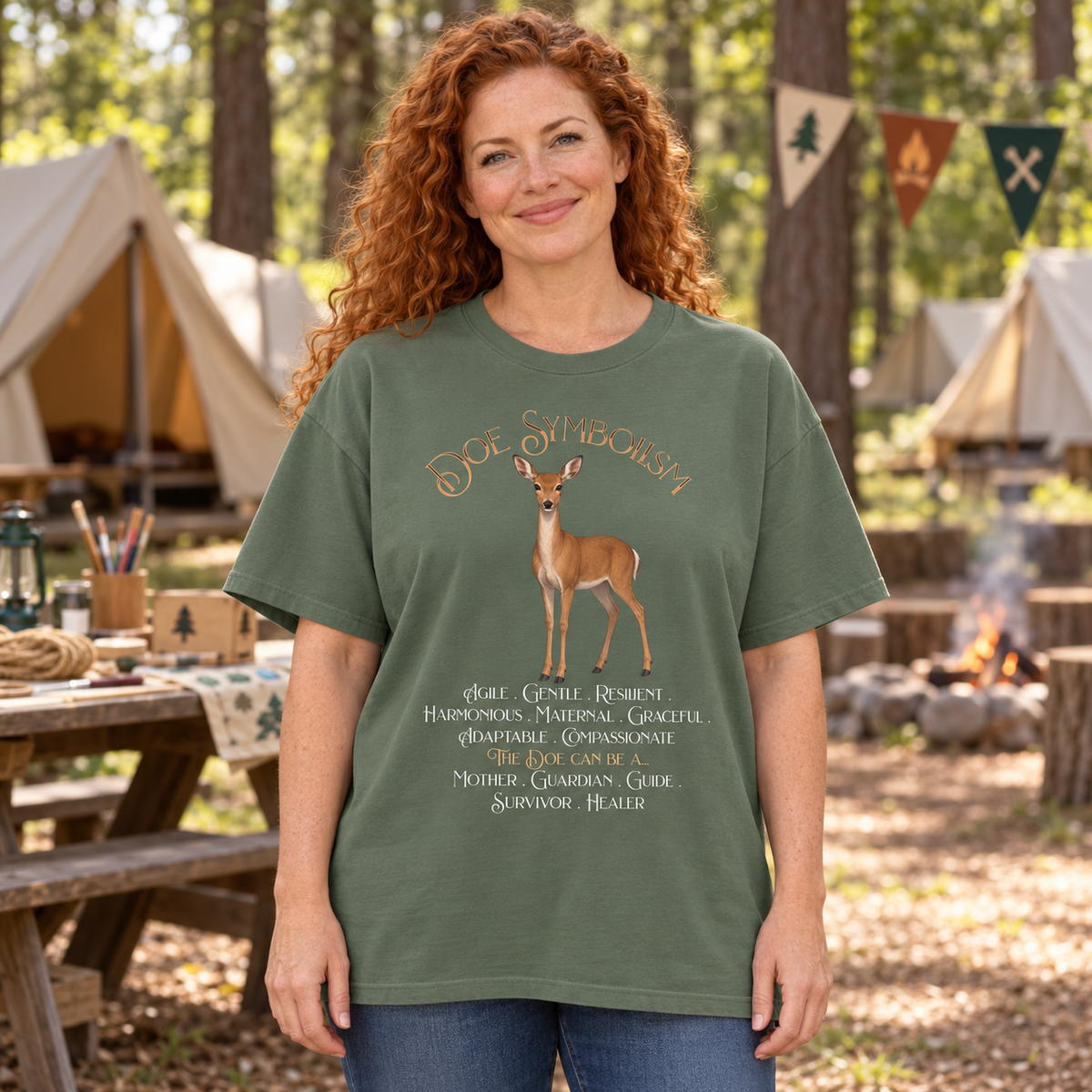 A woman with curly red hair stands outdoors in a guide campsite setting wearing an olive green graphic t-shirt with a deer illustration and text. The background shows tents, trees, and a fire pit.