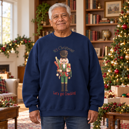 An older man wearing a navy blue crewneck sweatshirt with a Christmas nutcracker graphic and the text 'It's Christmas! Let's get Cracking'. He is standing in a cozy, decorated living room with a Christmas tree, boxes of ornaments, bookshelves, and holiday garlands.