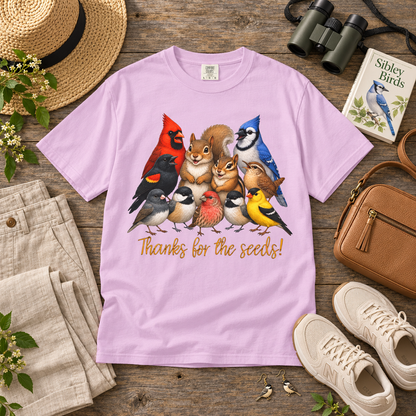A light purple crewneck t-shirt laid flat on a rustic wooden table, featuring a colorful graphic of various birds, a squirrel, and a chipmunk with the text 'Thanks for the seeds!'. The shirt is styled in a flat-lay arrangement with a straw hat, beige pants, binoculars, a book, a brown leather purse, and white sneakers.