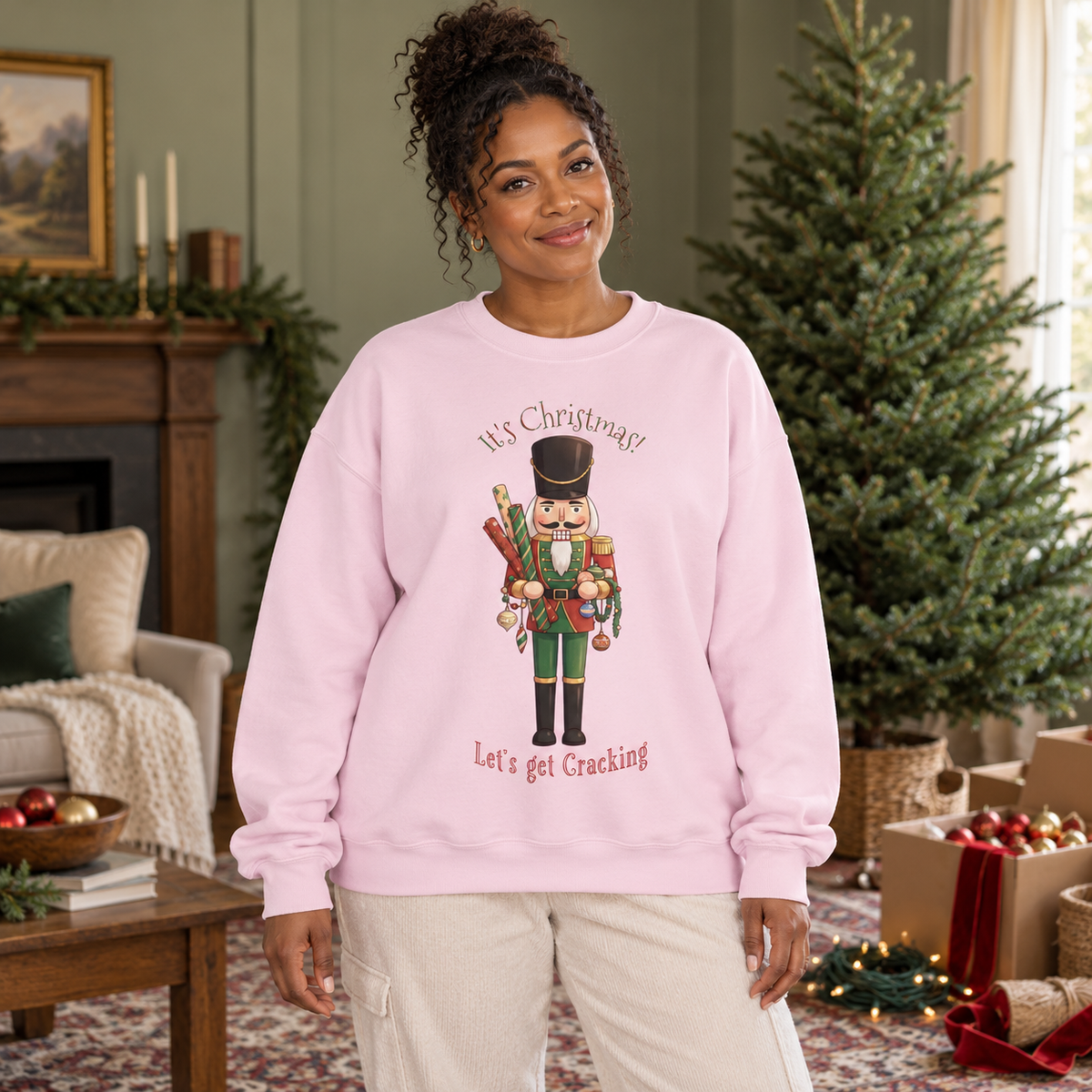 A woman wearing a light pink crewneck sweatshirt with a nutcracker graphic and the text 'It's Christmas! Let's get Cracking' in a cozy living room setting with boxes of oranaments, a Christmas tree, and fireplace in the background.