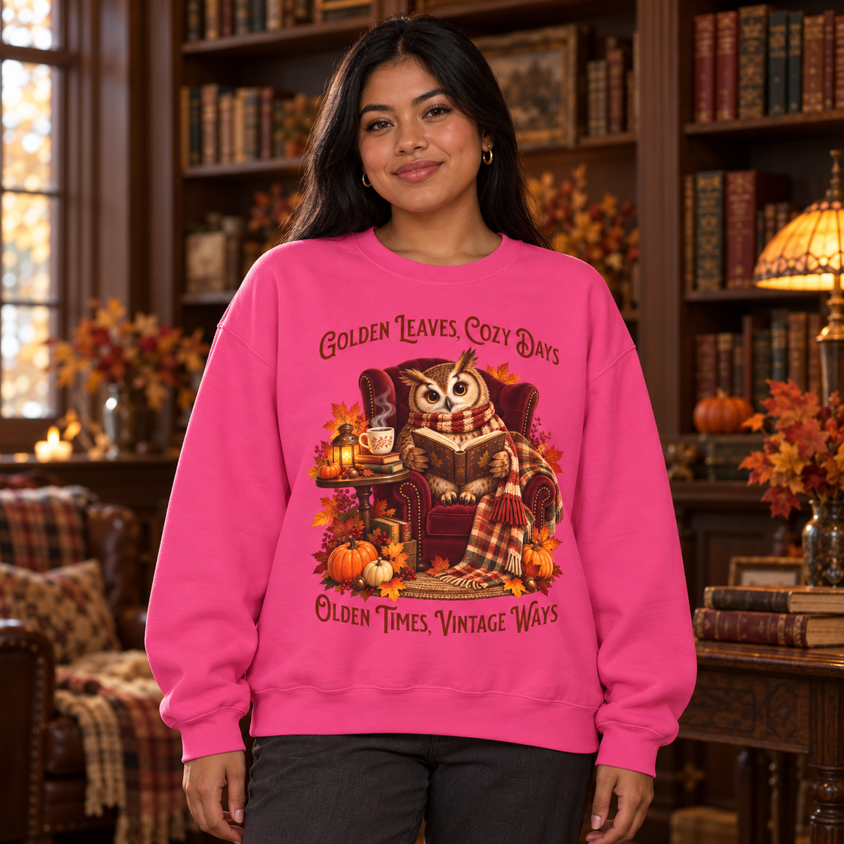A woman wearing a bright pink crewneck sweatshirt with a detailed autumn graphic of an owl curled up on a chair with a book and the text, 'Golden Leaves, Cozy Days, Olden Times, Vintage Ways'. The woman is standing in a cozy, warmly lit home library.