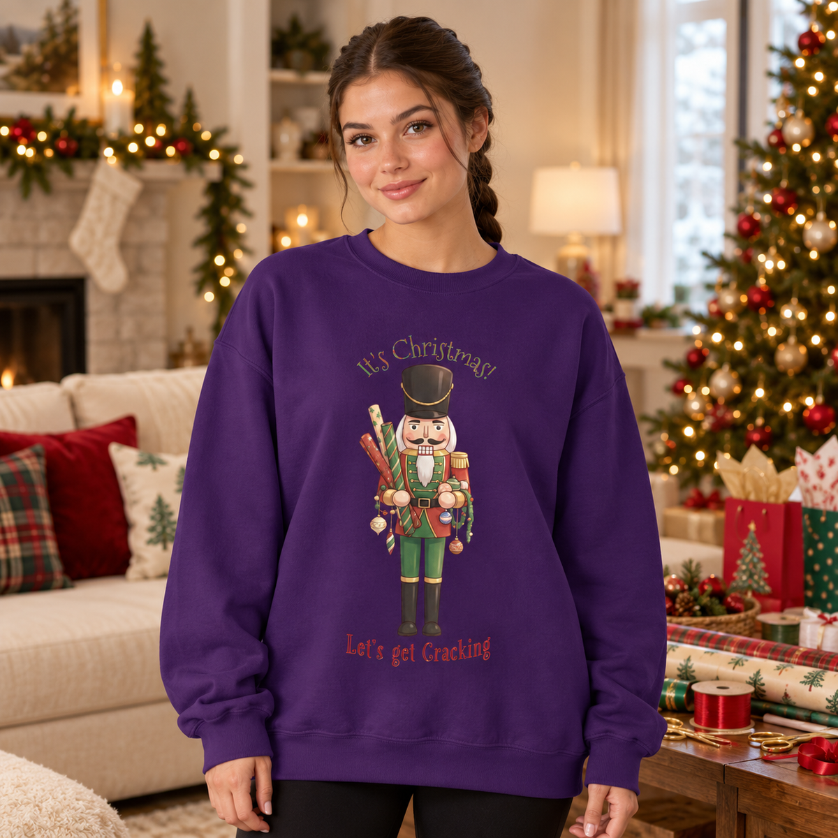 A woman wearing a purple crewneck sweatshirt with a nutcracker graphic and the text 'It's Christmas! Let's get Crackling' in a cozy, decorated living room with wrapping paper and a Christmas tree.